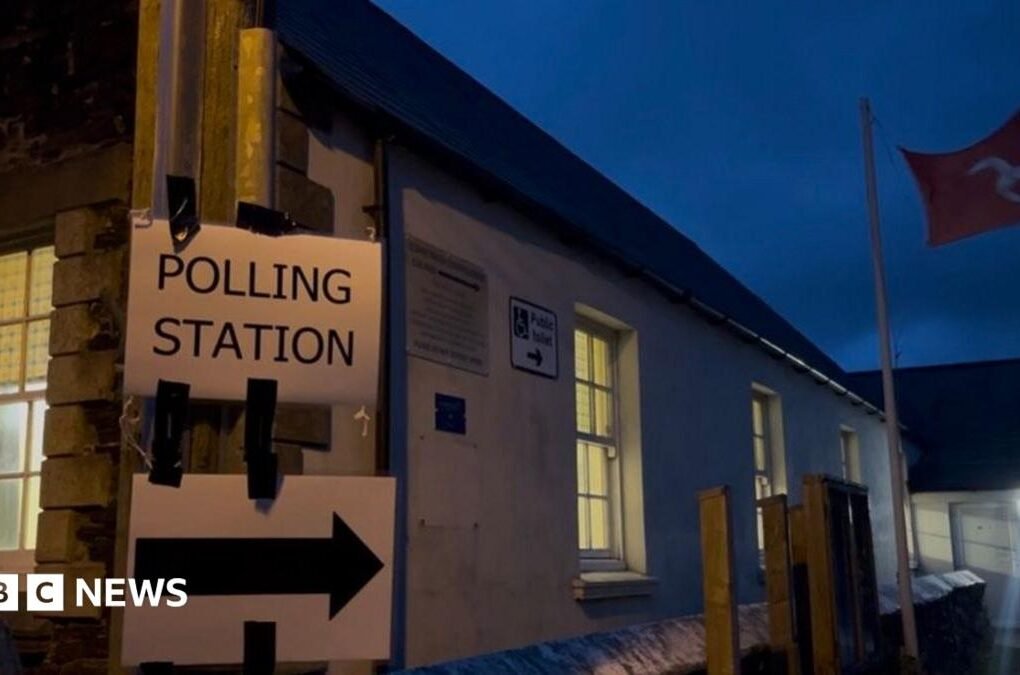 A sign reads polling station with an arrow, there is a Manx flag flying in the background.