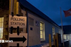 A sign reads polling station with an arrow, there is a Manx flag flying in the background.