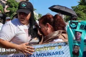 People take part in a march for peace in Culiacan, Mexico on 13 September 2026. A woman wearing a white T-shirt and a black baseball cap comforts another woman who is crying. The woman with the baseball cap is carrying a placard reading "help us find Jose Gonzalo Flores Molinares". In the background another protester is holding up another banner with the photos of missing people on it.