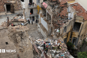 Site the blast showing the collapsed remains of the building, flanked on either side by heavily damaged properties